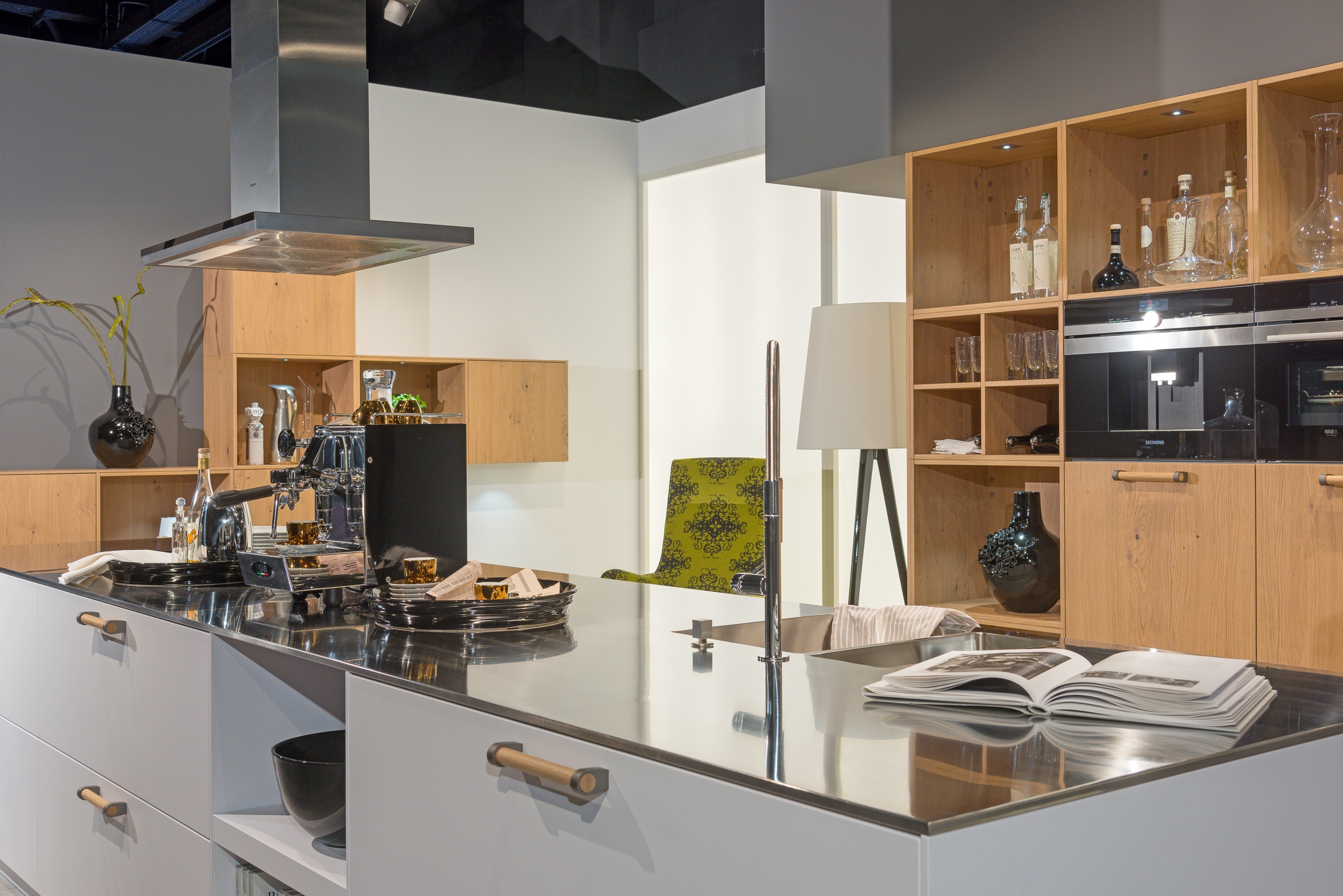 weiße Küche mit Kochinsel und Kochhaube, dazu Metallarbeitsplatte und Küchenschrank mit Einbauofen und offenen Regalelementen