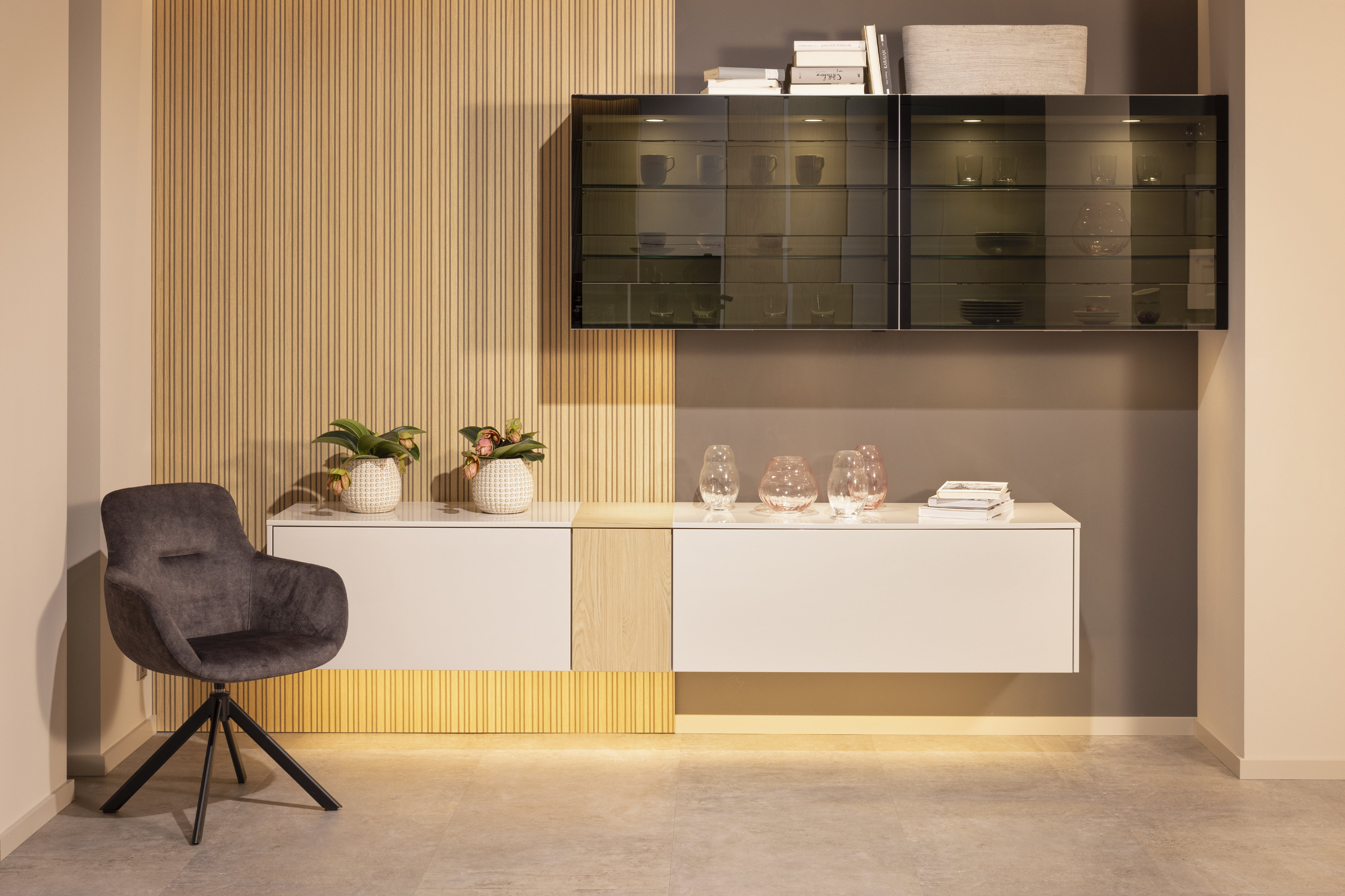 Wandmontiertes Lowboard in Weiss mit Holzeinsatz mittig schräg darüber schwarzer Hänge-Vitrinenschrank davor dunkelgrauer Samt-Polster-Drehstuhl